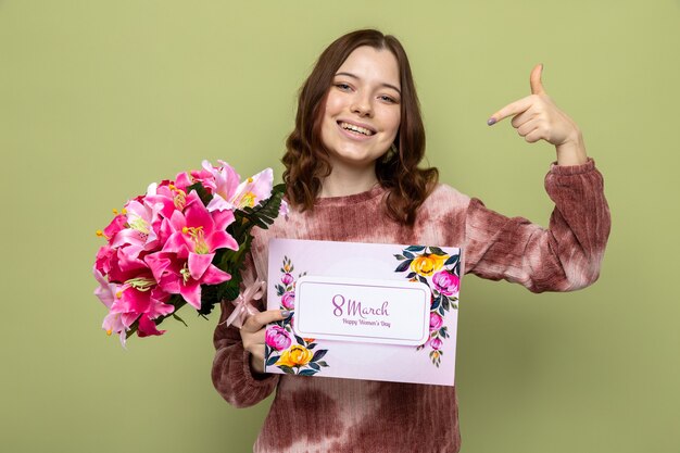 Smiling points at herself beautiful young girl on happy woman's day holding bouquet with postcard isolated on olive green wall