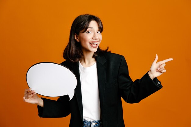 オレンジ色の背景に分離された黒いジャケットを着て吹き出し若い美しい女性を保持している側の笑顔のポイント