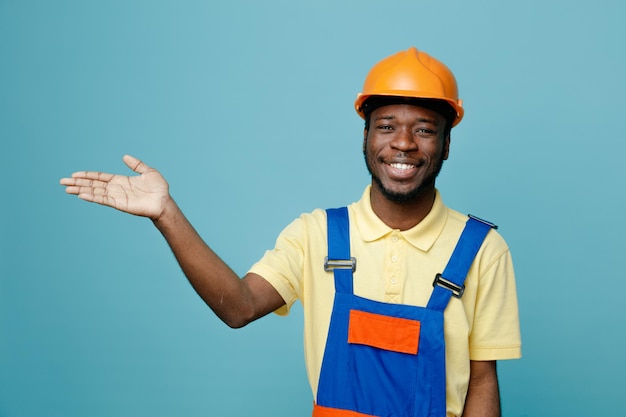 Smiling points at side young african american builder in uniform isolated on blue background
