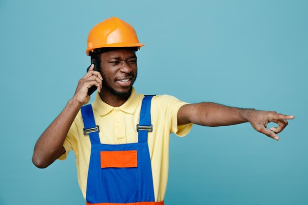 Улыбающийся молодой афро-американский строитель в форме разговаривает по телефону на синем фоне