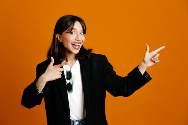 Smiling points at side young beautiful female wearing black jacket isolated on orange background