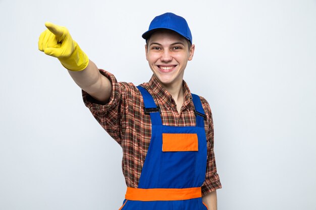 Smiling points at side young cleaning guy wearing uniform and cap with gloves 