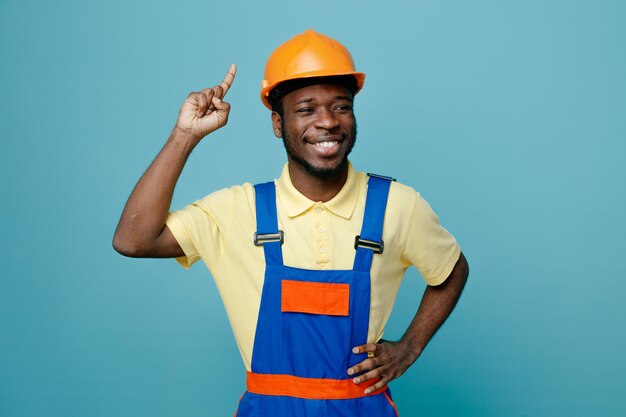 улыбаясь указывает на поднятую руку на бедра молодого афро-американского строителя в униформе, изолированного на синем фоне