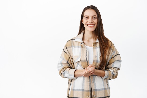 Smiling polite woman ready to help, wants to assist, hold hands together over chest and looking friendly at front, giving speech, standing against white wall