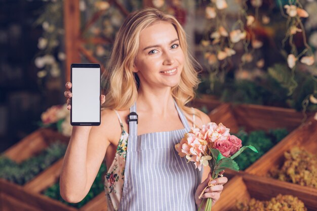 Smiling portrait of a blonde young woman holding bouquet in hand showing mobile phone