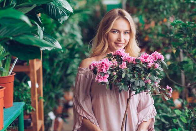 Smiling portrait of blonde young woman holding plant with pink flowers in the garden