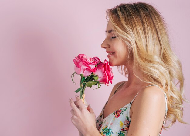 Smiling portrait of blonde young woman holding roses in hand against pink background