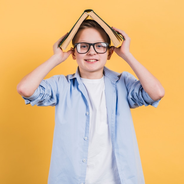 Smiling portrait of a boy holding an open book on his head standing against yellow backdrop