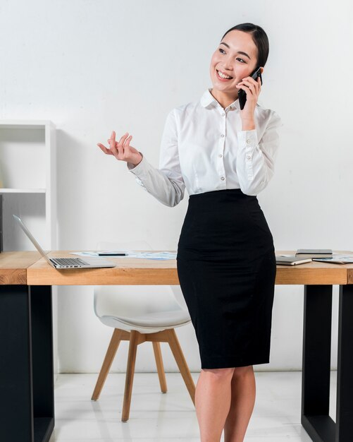 Smiling portrait of a businesswoman standing in front of desk talking on mobile phone gesturing