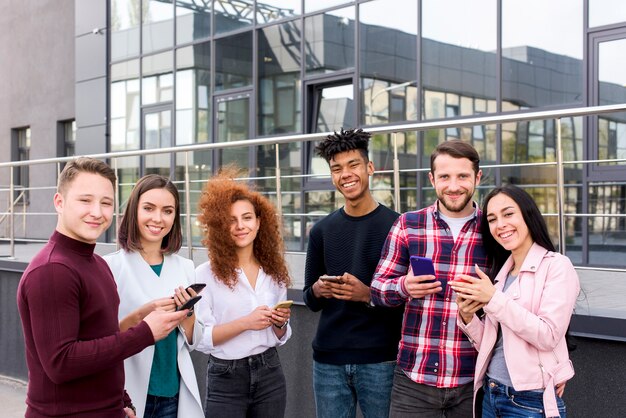 建物の外に立っているスマートフォンを使用して陽気な若い学生たちの笑顔の肖像画