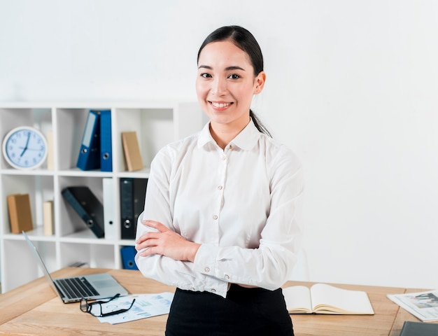 Smiling portrait of a confident young businesswoman standing in front of table looking to camera