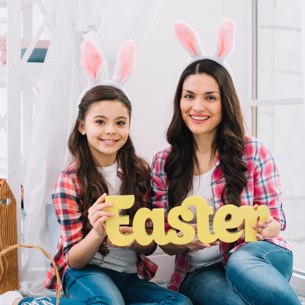 Smiling portrait of a daughter and mother with bunny ears holding easter text