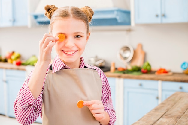 Smiling portrait of a girl covering her one eye with carrot in the kitchen