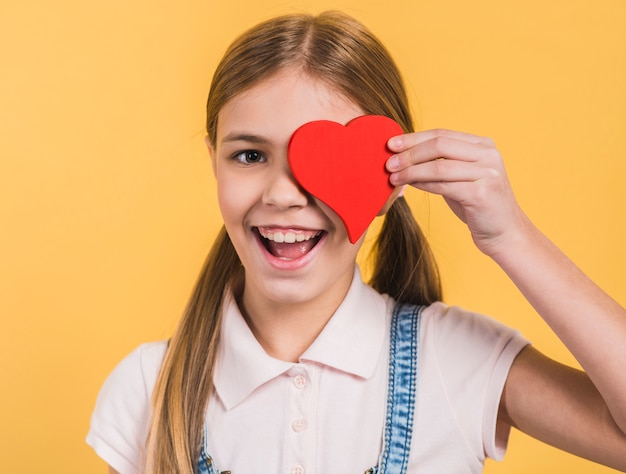 Smiling portrait of a girl holding red paper cut out heart shape in front of her eyes against yellow background