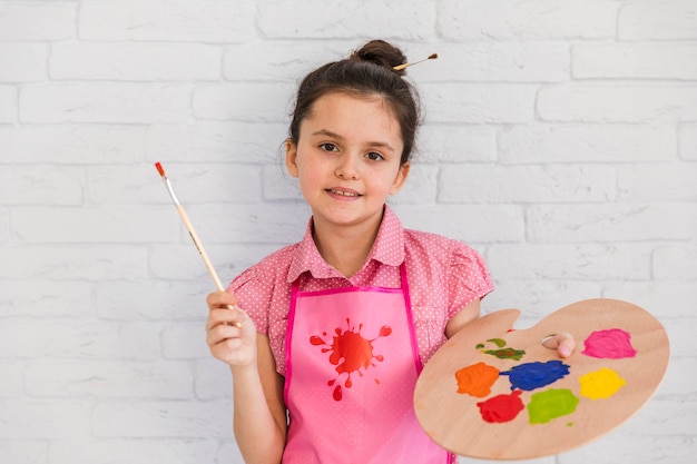 絵筆とカラフルなパレットを保持している白いレンガの壁に立っている女の子の肖像画を笑顔