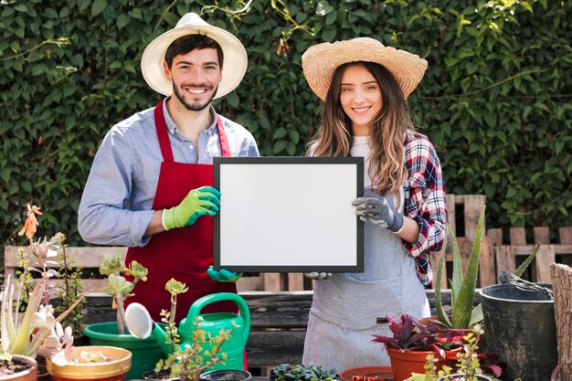 Усмехаясь портрет шляпы садовника мужчины и женщины нося показывая белую пустую рамку в саде