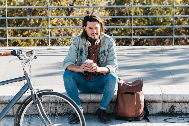 Smiling portrait of a man sitting on sidewalk with his backpack holding disposable coffee cup