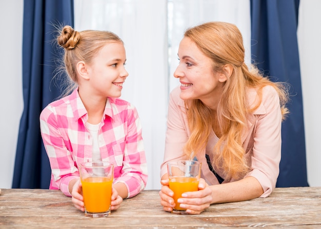 Smiling portrait of a mother and her daughter holding glass of juice looking at each other on wooden table