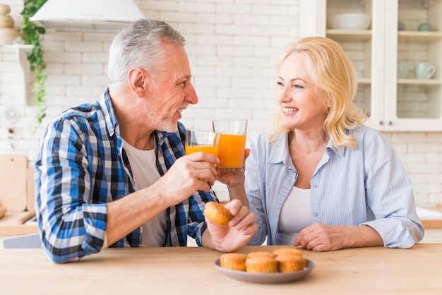 Smiling portrait of a senior couple toasting the glasses of juice and muffins on table in the kitchen