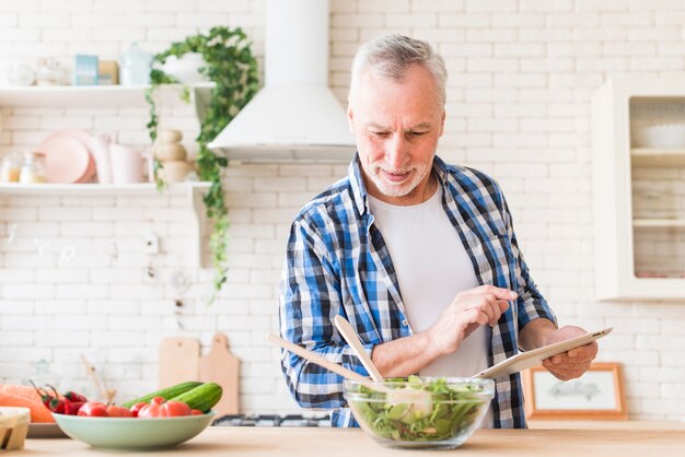 Улыбаясь портрет старшего человека, приготовление пищи с использованием цифрового планшета на кухне