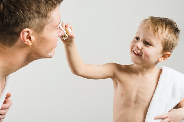 Smiling portrait of a shirtless blonde boy applying shaving foam on his father's nose against gray background