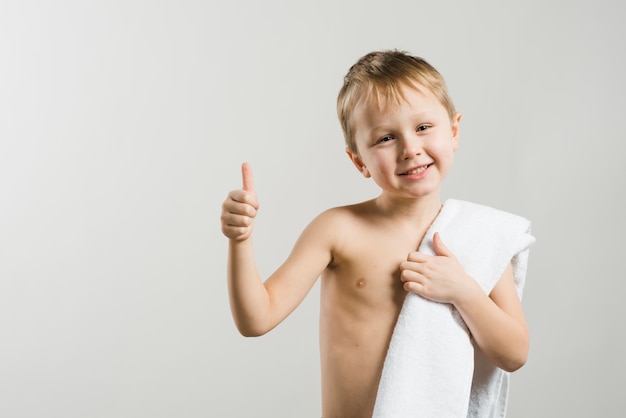 Улыбающийся портрет без рубашки белокурый мальчик с белым полотенцем на плече, показывая большой палец вверх знак на сером фоне