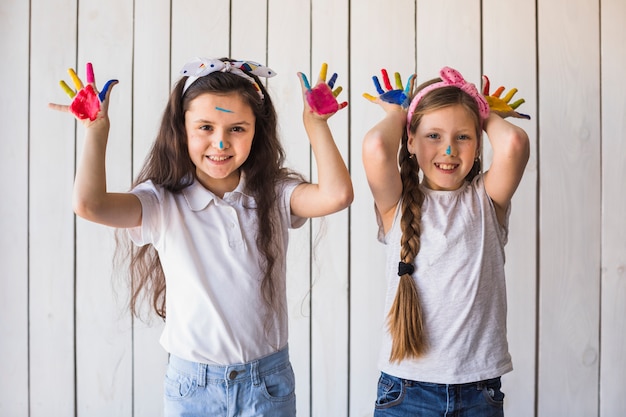 Foto gratuita un ritratto sorridente di due ragazze che mostrano le mani dipinte variopinte che stanno contro la parete di legno