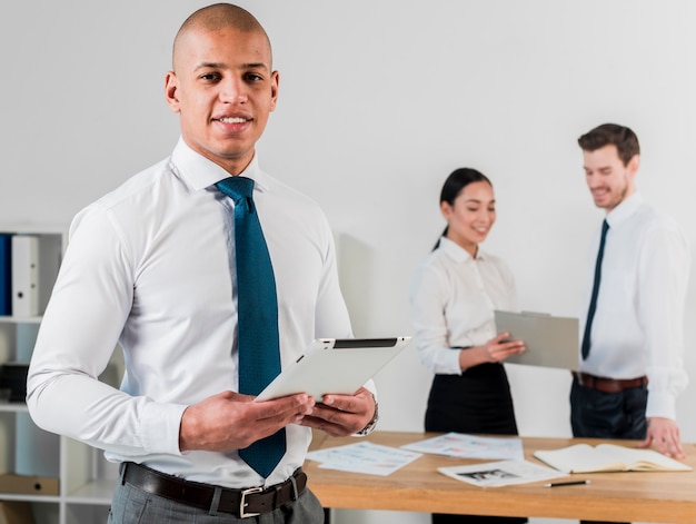 Smiling portrait of a young businessman holding digital tablet in hand and his colleague working at background