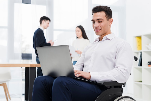Smiling portrait of a young businessman sitting on wheelchair using laptop with his colleague at background
