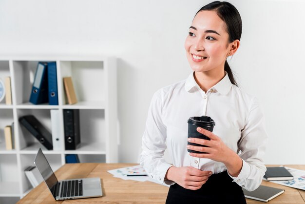 使い捨てのコーヒーカップを手で押しテーブルの前に立っている若い実業家の肖像画を笑顔