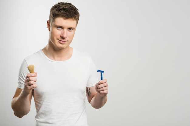 Smiling portrait of a young man holding razor and shaving brush in hands