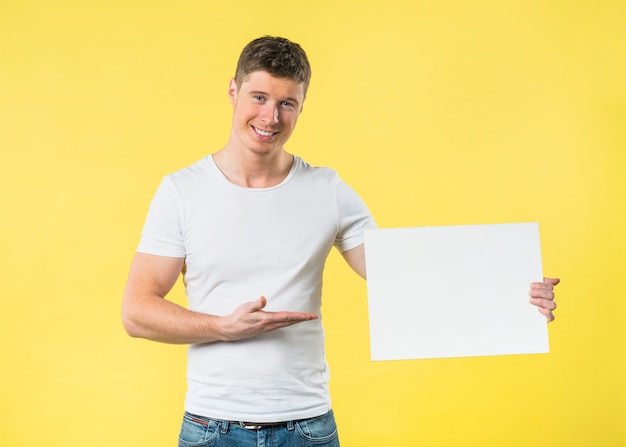 Smiling portrait of a young man presenting something on white blank card against yellow backdrop