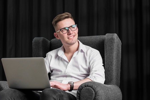 Smiling portrait of a young man sitting on arm chair using digital tablet looking away