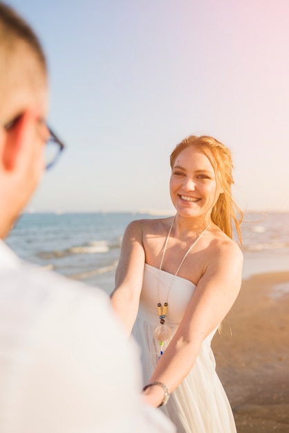 Усмехаясь портрет молодой женщины держа руки его парня наслаждаясь на пляже