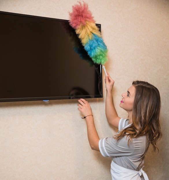 Free photo smiling portrait of a young woman using duster to clean the television screen on wall