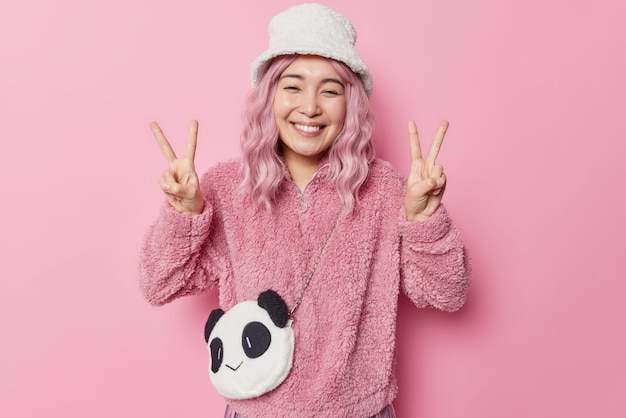 Smiling positive Asian woman makes peace gesture with both hands being in good mood enjoys life wears warm fur jacket and fashionable panama carries small bag gestures against pink background