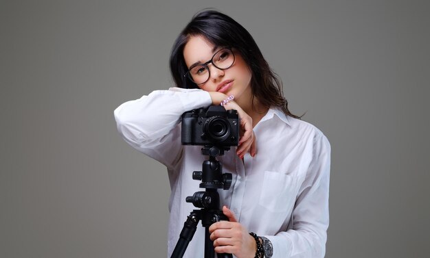 안경을 쓰고 전문 사진 카메라로 사진을 찍고 있는 웃고 있는 긍정적인 갈색 머리 여성. 회색 배경에 고립.