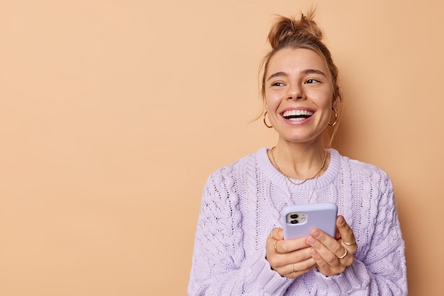 Smiling positive European woman with combed hair uses cellphone enjoys modern technologies holds mobile phone has chat online wears casual sweater isolated over beige background blank space