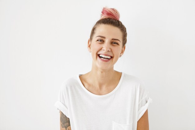 Free photo smiling positive female with attractive look, wearing loose white t-shirt, posing against white blank wall. happy woman with hair bun showing positive emotions after receiving pleasant compliment