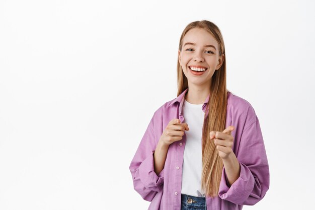 Smiling positive girl, pointing fingers at front, inviting, picking someone, praising your efforts, standing against white wall