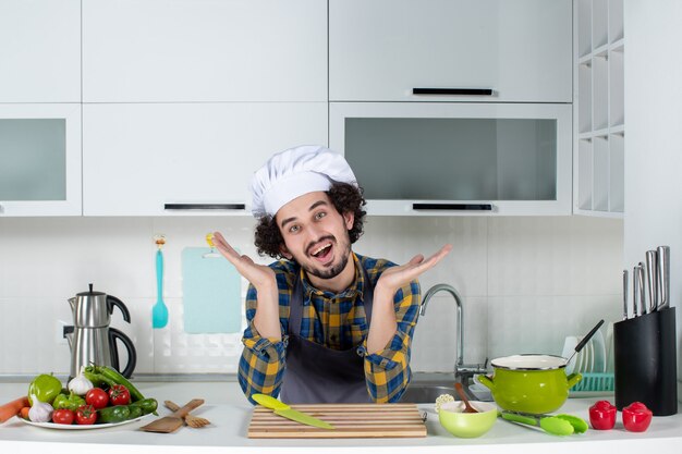 Улыбающийся и позитивный шеф-повар со свежими овощами позирует на белой кухне