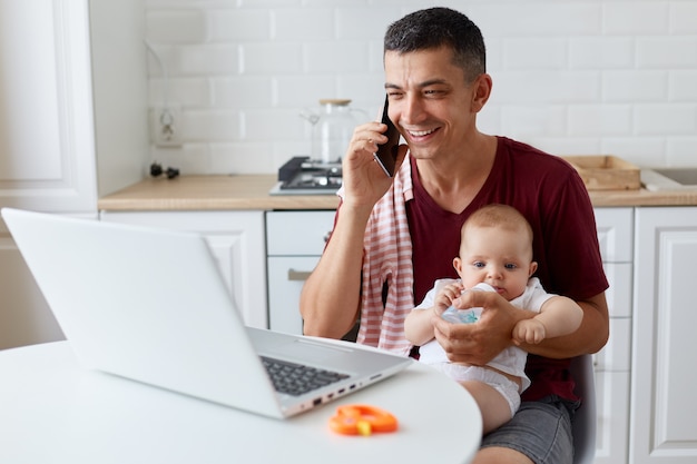 Smiling positive man wearing maroon casual t shirt sitting in kitchen in front of laptop computer and talking phone, giving water from bottle for his daughter, working online.