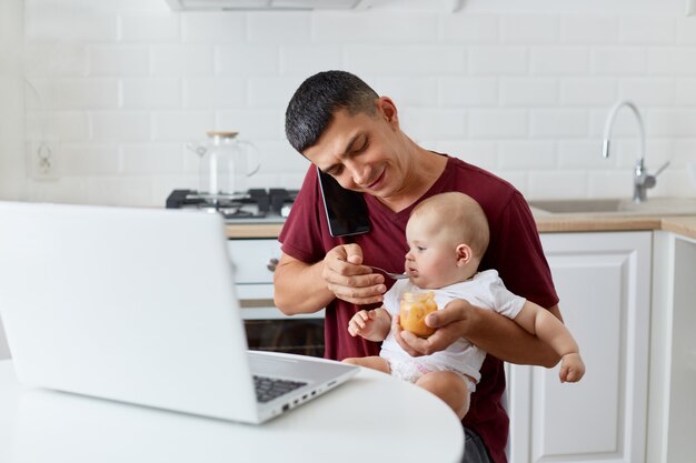 Smiling positive man wearing maroon casual t shirt talking phone while feeding little daughter or son with fruit puree, sitting at table in kitchen in front notebook.