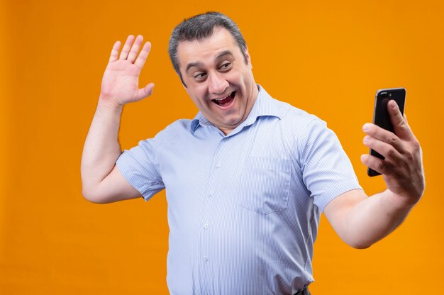 Smiling and positive middle age man in blue striped shirtwear headphones waving hand say hello as talking on video-call using his smartphone while standing on an orange background