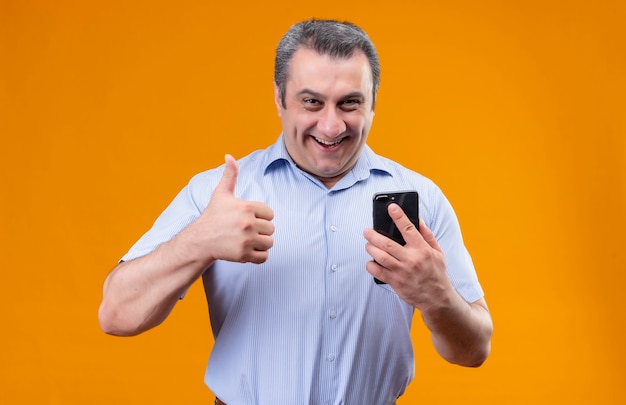 携帯電話を押しながらオレンジ色の背景の上に立っている間親指を現して青いストリップシャツを着て笑顔で肯定的な中年男