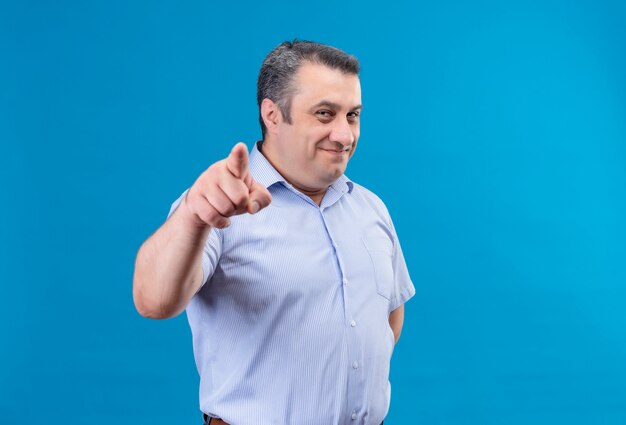 青色の背景にカメラで人差し指を指している青い縞模様のシャツを着ている笑顔の中年男性