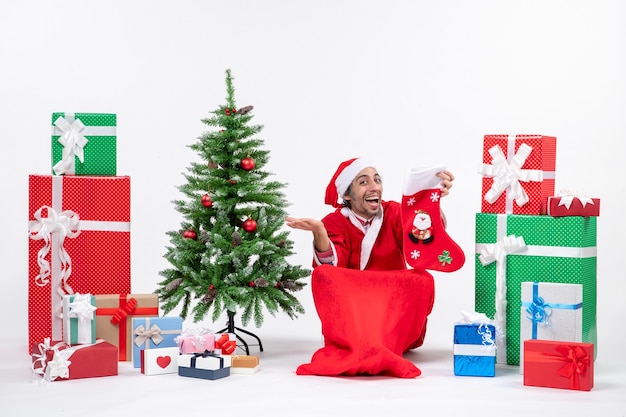 Smiling positive santa claus sitting on the ground and holding christmas sock near gifts and decorated new year tree on white background