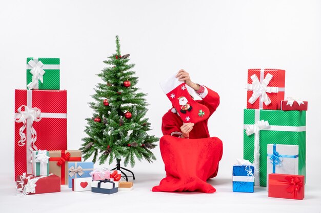 Улыбающийся позитивный санта-клаус сидит на земле и поднимает рождественский носок к лицу возле подарков и украшает новогоднюю елку на белом фоне