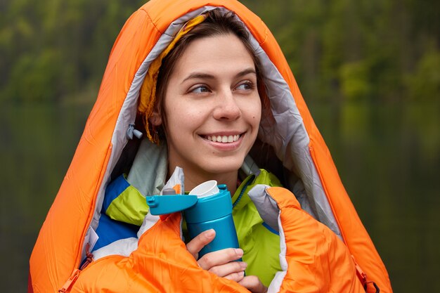 Smiling positive young female traveler wrapped in sleeping bag, warms with hot drink during cold autumn day