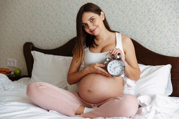 Улыбается беременная женщина в помещении дома сидит на кровати
